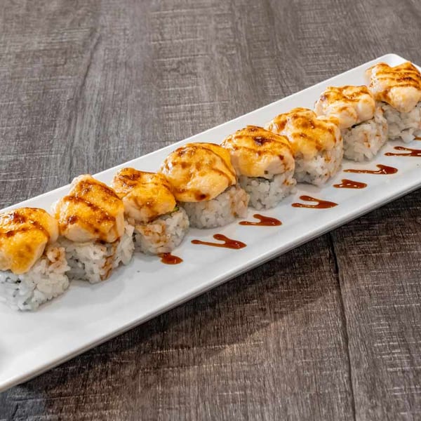 A white rectangular plate holds a row of eight baked sushi rolls with sauce drizzles on a wooden table.