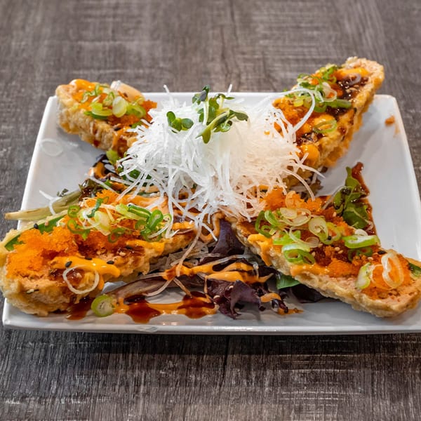Four crispy sushi rice triangles topped with orange roe, green onions, and sauces on a white plate.